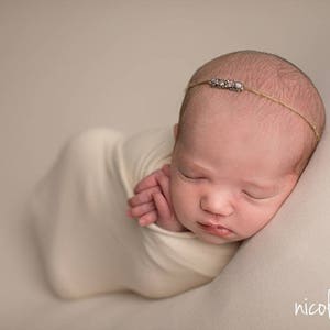 May include: A newborn baby girl is swaddled in a white blanket and is sleeping peacefully. She is wearing a delicate headband with rhinestones.