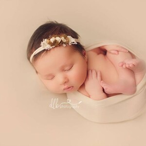 May include: A newborn baby girl is sleeping soundly in a white swaddle. She is wearing a white headband with brown and white flowers. The baby is lying on a light pink background.