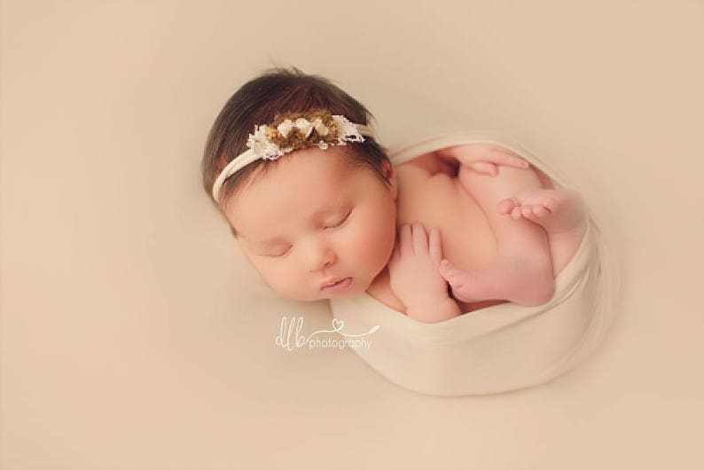 May include: A newborn baby girl is sleeping soundly in a white swaddle. She is wearing a white headband with brown and white flowers. The baby is lying on a light pink background.