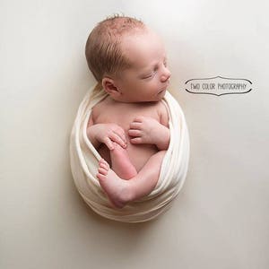 May include: A newborn baby is swaddled in a white fabric and is sleeping peacefully. The baby is lying on a white background. The image is taken from above.