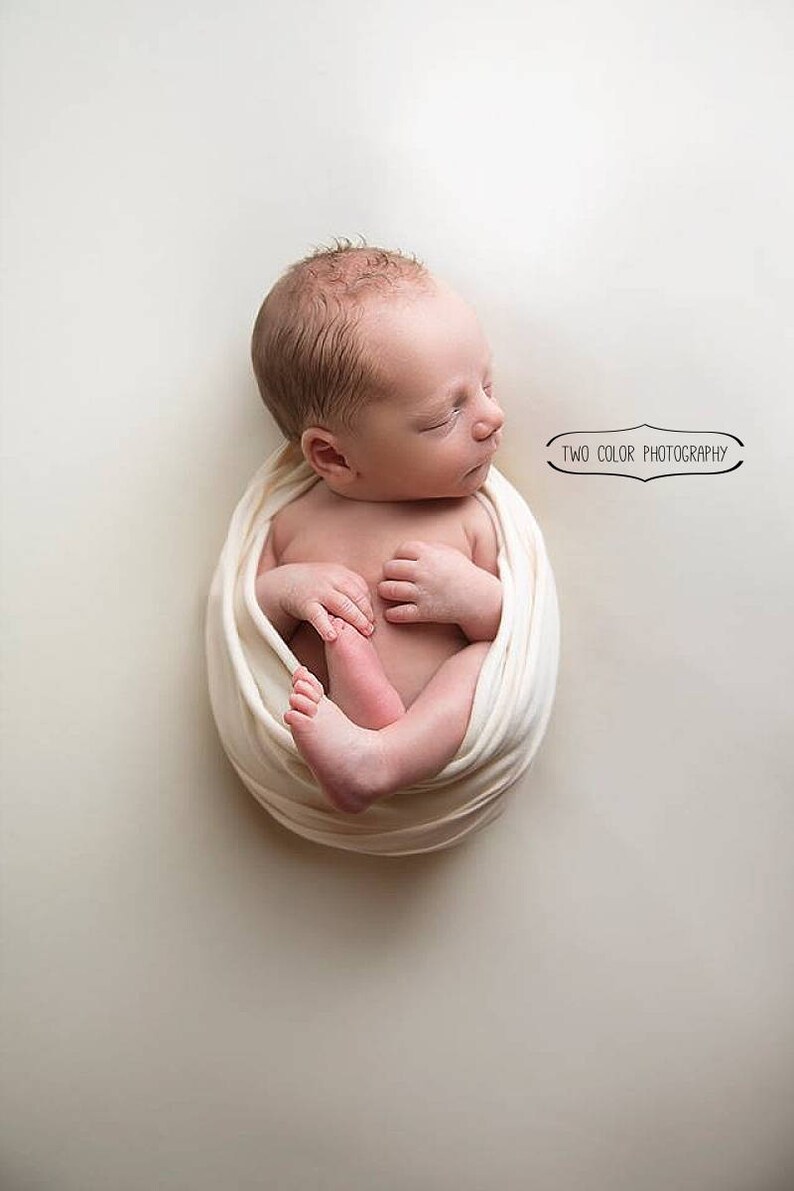 May include: A newborn baby is swaddled in a white fabric and is sleeping peacefully. The baby is lying on a white background. The image is taken from above.