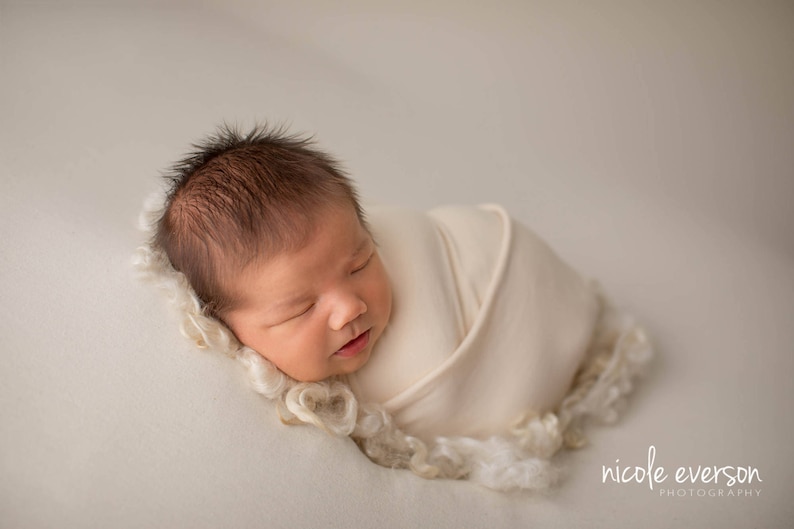 May include: A newborn baby is sleeping soundly, wrapped in a white swaddle blanket. The baby has dark hair and is lying on a white background.