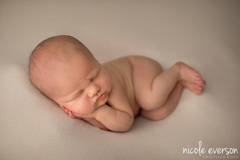 May include: A newborn baby is sleeping on a white blanket. The baby is lying on its side with its head resting on its arm. The baby's eyes are closed and its mouth is slightly open.