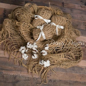 May include: A brown burlap fabric backdrop with white cotton balls and a stack of brown burlap squares tied with white ribbon. The fabric is arranged in a loose, flowing style, creating a rustic and natural feel.