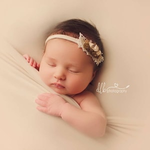 May include: A newborn baby girl sleeping peacefully in a white swaddle. She is wearing a white headband with a floral design.