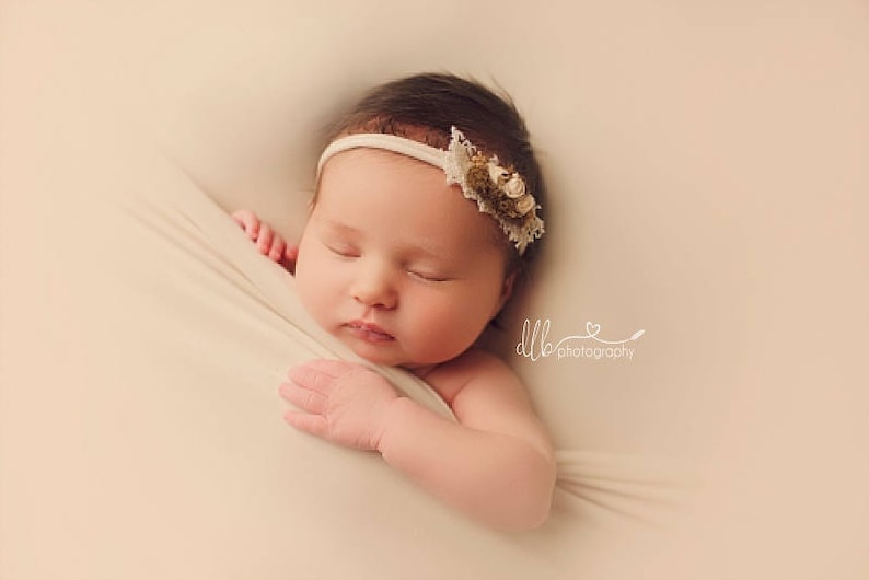 May include: A newborn baby girl sleeping peacefully in a white swaddle. She is wearing a white headband with a floral design.