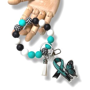 May include: A beaded keychain bracelet with a variety of beads in black, white, teal, and checkered patterns. The keychain includes a white tassel and a charm with a teal ribbon and butterfly design.