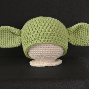 May include: Green crocheted hat with large ears, resembling a popular fictional character. The hat is on a white crocheted form.