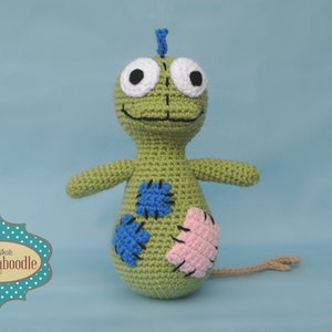 May include: A green crocheted toy with a long tail, blue and pink patches, and large white eyes. The toy is smiling and has a blue antenna on its head.