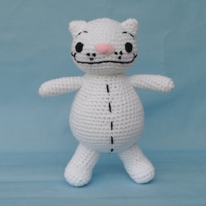 Könnte beinhalten: Weißes gehäkeltes Katzen-Plüschtier mit schwarzen Augen und einer rosa Nase. Die Katze hat ein gesticktes Lächeln und eine schwarze Linie auf dem Bauch.