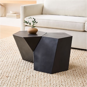 May include: Two black geometric side tables with a faceted design. The tables are nested together and a small brown vase with a green plant is on the top table.