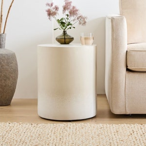 May include: A white round side table with a subtle ombre effect. The table is sitting on a woven rug in front of a beige sofa.