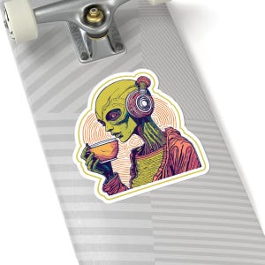 Op de afbeelding: Een sticker met een alien met groene huid en een gele kop, met een koptelefoon en een kopje. De illustratie is kleurrijk en gestileerd, met een retro-ontwerp.
