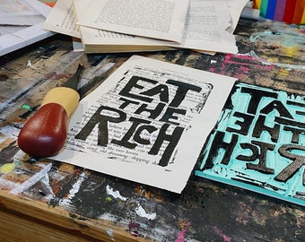May include: Two wooden printing blocks with the phrase "Eat the Rich" printed in black ink on a turquoise background. The blocks are sitting on a wooden table with paint splatters.
