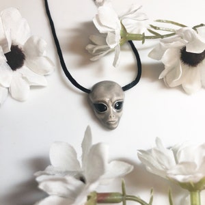 May include: A grey alien face pendant necklace with black eyes on a black cord. The pendant is surrounded by white flowers.