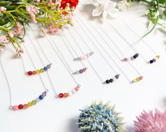 May include: A set of seven silver necklaces with different colored gemstone beads. Each necklace has a row of five to seven beads in a rainbow color pattern. The necklaces are displayed on a white surface with flowers.