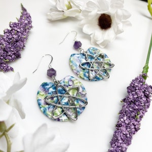 May include: A pair of earrings with a silver atom charm and a purple amethyst bead. The earrings are made with a floral pattern in shades of blue, green, and white.