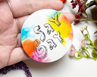 Può includere: Un adesivo rotondo con uno sfondo ad acquerello in tonalità di blu, arancione, giallo e rosa. La parola "Say Gay" è scritta in inchiostro nero sull'adesivo.