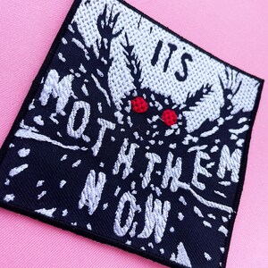 Puede incluir: Parche bordado negro con puntadas blancas que representa una polilla con ojos rojos y el texto "IT'S NOT THEM NOW".
