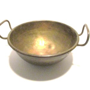 May include: A round, metallic bowl with a dull gold-toned finish and a hammered texture. The bowl features two curved, upright handles attached to opposite sides of the rim, providing a symmetrical appearance for this open-top kitchen vessel.