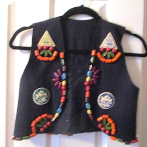 May include: A navy blue vest with colorful wooden beads and fabric appliques. The vest has a triangular applique on each shoulder and a circular applique on each side.