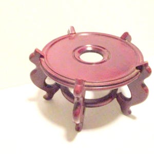 May include: A small, round, burgundy-colored wooden stand with a central hole. The stand has three curved legs and decorative cutouts. It is designed to hold a small object, such as a vase or figurine. The stand is approximately 4 inches in diameter.