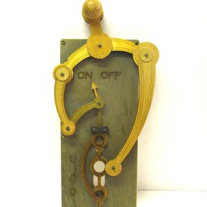 May include: A rectangular wooden wall plaque featuring a mechanical lever mechanism with a yellow handle. The plaque is painted a muted green and displays the text ON OFF with an arrow indicator. The design includes curved wooden arms and circular joints.