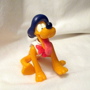 May include: A yellow plastic Pluto figurine with a blue hat, black nose, and pink tongue. The dog is wearing a white shirt with a red bow. The figurine is in a sitting position.