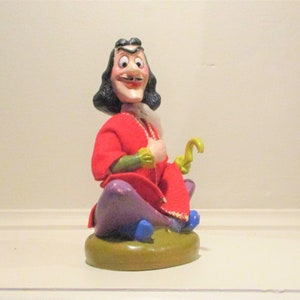 May include: A figurine of Captain Hook from Peter Pan. The figure is seated with a red coat, purple pants, and a yellow hook. The base is green with blue accents. The character has a white face, black hair, and a mustache.