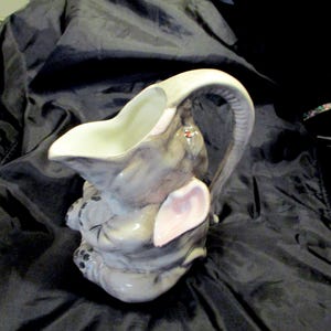 May include: A ceramic elephant-shaped pitcher with a white interior and a gray exterior. The pitcher has a curved handle that resembles an elephant's trunk. The elephant's ears are pink, and the pitcher is sitting on a dark surface.