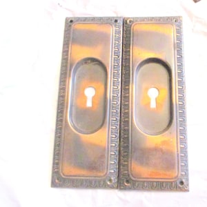 May include: Two antique bronze door handle plates. Each plate has an oval keyhole and a decorative border. The bronze has a patina with shades of brown and gold. The plates are rectangular and have screw holes.