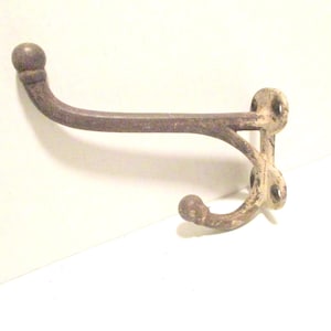 May include: A vintage-style, double-hook wall mount. The metal hook has a distressed, aged appearance with a rounded top and two curved hooks. The color is a mix of brown and off-white, suggesting an antique finish.