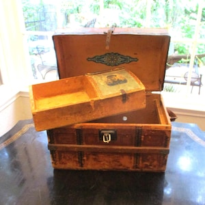 May include: A vintage brown wooden trunk with a hinged lid and a metal latch. Inside, a removable wooden tray rests on the top edges, featuring a small rectangular image of a person on its surface. The interior lid displays a decorative blue design.