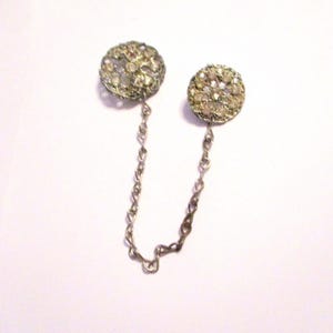 May include: A vintage silver-toned collar pin with two round, ornate, rhinestone-studded accents connected by a delicate chain. The rhinestones are clear and sparkling, set in a filigree design.