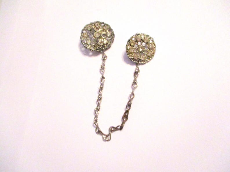 May include: A vintage silver-toned collar pin with two round, ornate, rhinestone-studded accents connected by a delicate chain. The rhinestones are clear and sparkling, set in a filigree design.