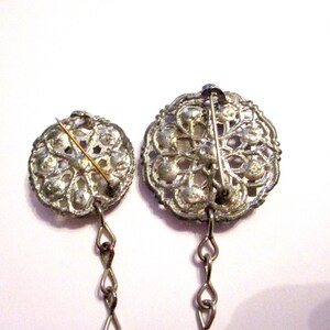 May include: Two vintage silver-toned brooches with intricate floral designs. Each brooch features a pin on the back and a delicate chain. The brooches are set against a white background, highlighting their ornate details.