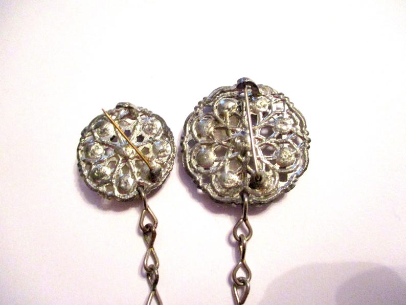 May include: Two vintage silver-toned brooches with intricate floral designs. Each brooch features a pin on the back and a delicate chain. The brooches are set against a white background, highlighting their ornate details.