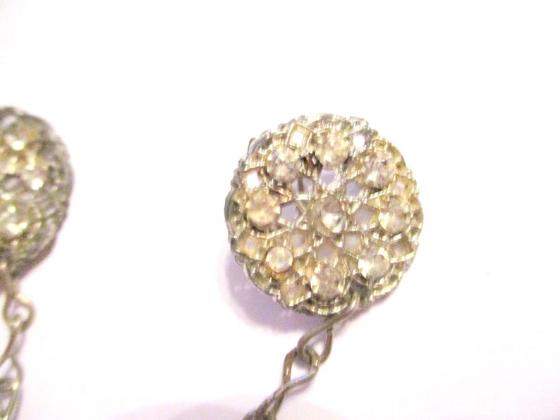 May include: Close-up of a pair of vintage earrings. Each earring features a round, ornate design encrusted with small, clear rhinestones. The earrings have a silver-toned metal setting and a delicate chain.