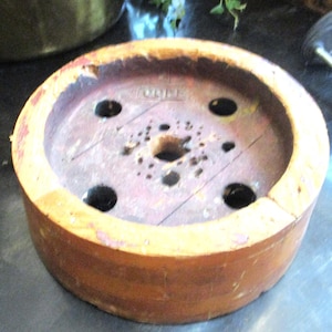 May include: A circular wooden hat block with a weathered orange finish and a recessed center. The interior surface is dark brown, featuring a central hole surrounded by four larger circular openings and several smaller, scattered drill holes.