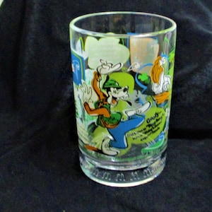 May include: A clear glass featuring a colorful cartoon design of Goofy in a green hat and blue pants. The glass has a wide base and a cylindrical shape, with additional cartoon characters and text. The glass is likely a collectible item.