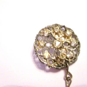 May include: A vintage brooch with a round, openwork design. The brooch features a gold-toned metal frame and is adorned with numerous clear and amber-colored rhinestones. A small chain hangs from the bottom.