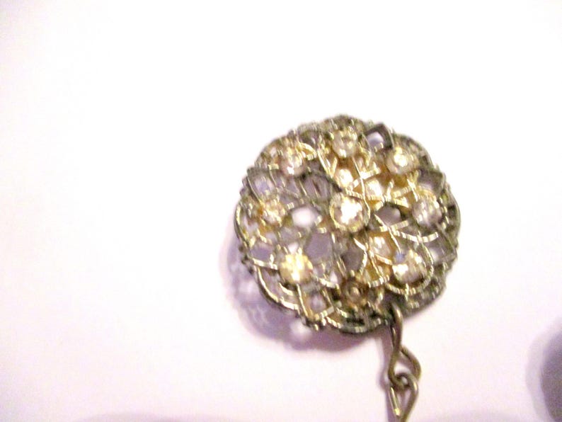 May include: A vintage brooch with a round, openwork design. The brooch features a gold-toned metal frame and is adorned with numerous clear and amber-colored rhinestones. A small chain hangs from the bottom.