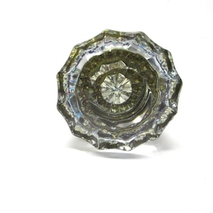 May include: A clear, faceted glass door knob with a multi-sided, starburst-like geometric pattern radiating from the center. The knob features a reflective, transparent surface with distinct ridges and a circular, scalloped outer edge design.