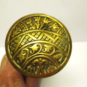 May include: A round brass door knob featuring an intricate embossed design with floral motifs, curved lines, and geometric patterns. The metal surface has a darkened patina in the recessed areas, highlighting the detailed relief work.