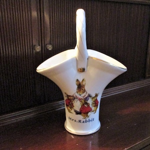 May include: White ceramic basket with a handle, featuring a Mrs. Rabbit illustration and gold trim. The basket has a flared shape and a curved handle. The illustration depicts a rabbit and her children in red and purple clothing.