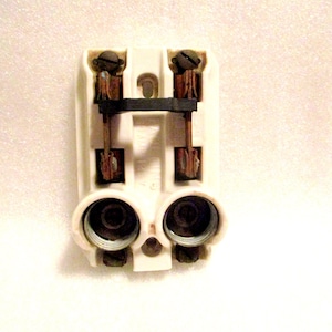 May include: A vintage electrical component with a cream-colored plastic housing. It features two screw terminals, copper wiring, and two circular receptacles. The design suggests it is a fuse holder or a similar electrical device.