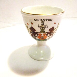 May include: White ceramic egg cup with a gold rim and a colorful illustration of the coat of arms of Southampton, England. The cup has a short stem and a round base.