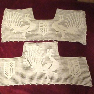 May include: Off-white, hand-crocheted lace pieces, possibly for a garment. The pieces feature a detailed design with floral and geometric patterns. The crochet work is visible, with a textured appearance. The pieces are displayed on a dark red surface.