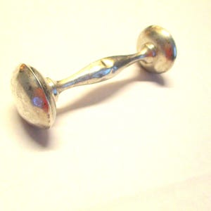 May include: A small, silver-colored dumbbell with rounded ends and a twisted handle. The dumbbell is lying on a white surface, casting a shadow. The object appears to be made of metal.