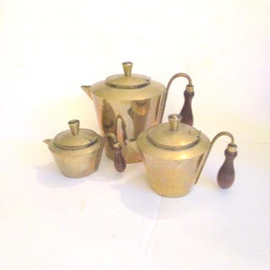 May include: A vintage tea set with a brass finish, including a teapot, a creamer, and a sugar bowl. Each piece has a dark wooden handle and a lid with a small knob. The set is displayed on a white surface.